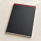 A black textured notebook with a dark red top band and the brand name 'INKFORGE' debossed near the bottom, resting on a light beige fabric background.