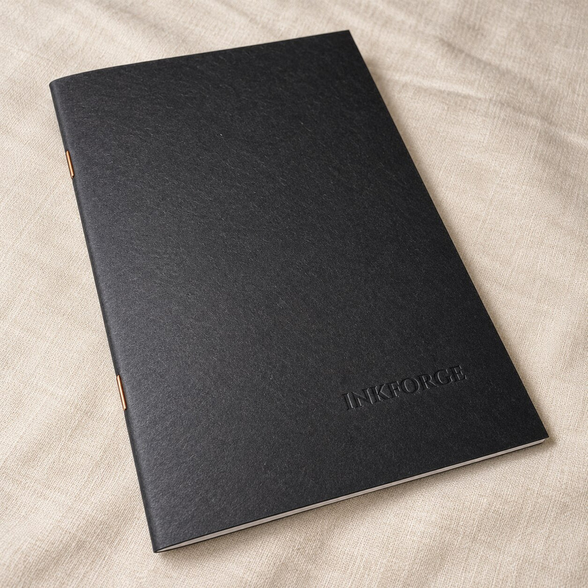 A black paperback notebook with a blind debossed Inkforge logo in the bottom right corner, resting on a beige linen fabric background.