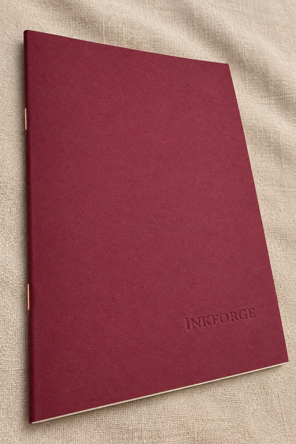 A paperback notebook with a burgundy cover, lying on a beige linen fabric background. The brand name Inkforge is blind debossed in the bottom right corner.