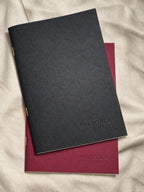 Two paperback notebooks, one in black and the other in deep burgundy, both blind embossed with the brand name Inkforge and stacked on a beige fabric background.